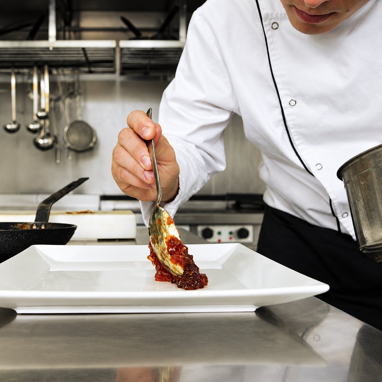 Cuisinier en veste blanche dressant une assiette avec une cuillère dans une cuisine professionnelle en inox