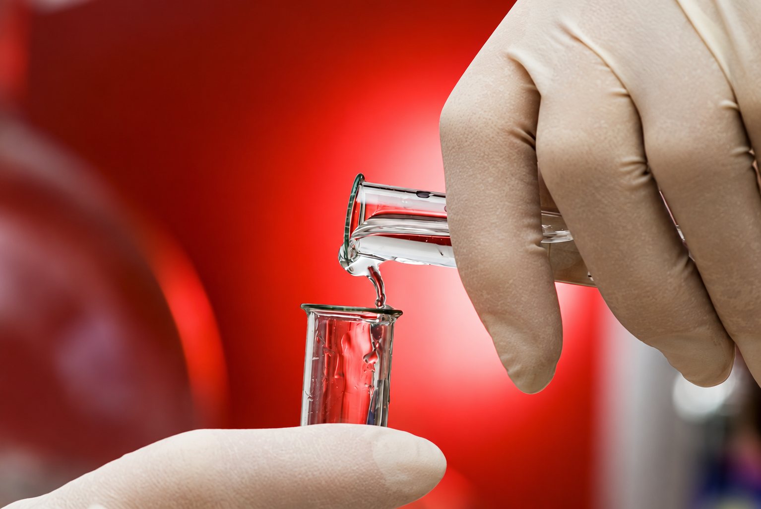 Main gantée versant un liquide transparent d’une éprouvette dans un petit bécher, sur fond rouge flou